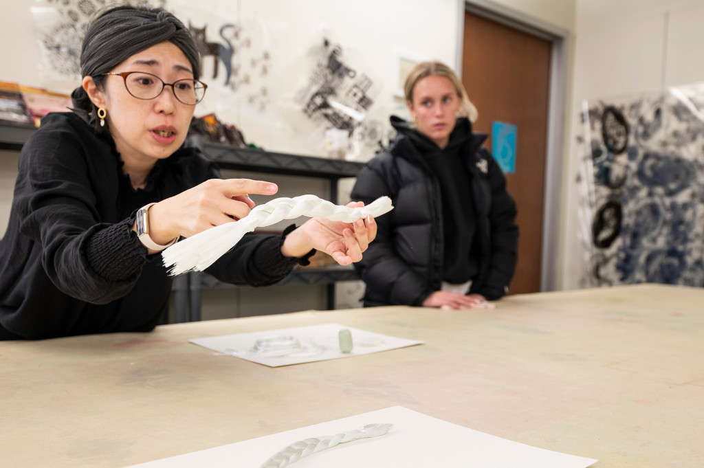 Machiko Ito leans across a large table and displays a work of braided, kiln-fired fiberglass.