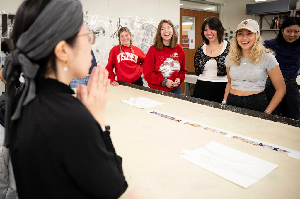 Machiko Ito gestures with her palms together as she speaks across a large wooden table to a class of undergraduate students.
