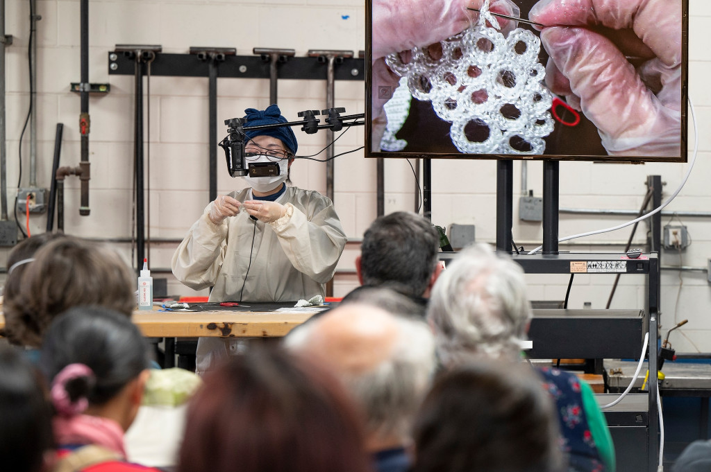 Machiko Ito stands in the Glass Lab in front of a seated audience. She works at a crocheted fiberglass circle, enlarged for the audience on a screen next to her work table.