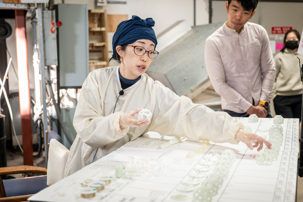 In the Glass Lab, Machiko Ito wears a protective smock and handles finished glass pieces selected from a table.