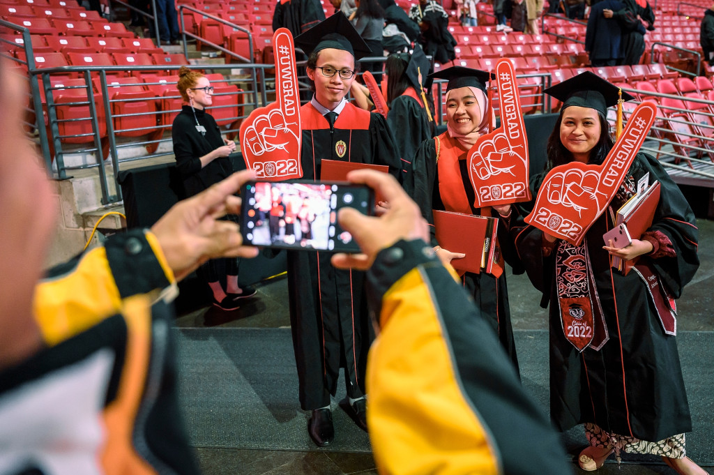 People in graduation gowns smile for a photo being taken by someone in the foreground with a cell phone.