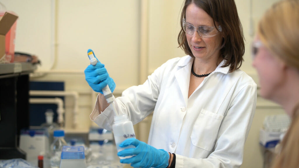 In a lab setting, Christy Remucal wears a white lab coat, protective glasses and gloves while dipping a pipette into a sample bottle.