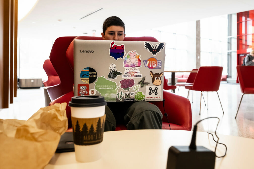 A person sits in a red chair, with a coffee on the table in front of them, and looks at a laptop.