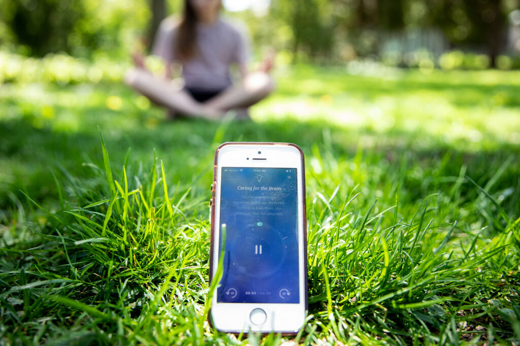 An iPhone resting in the grass shows the Healthy Minds Program app in use while a person meditates in lotus pose.