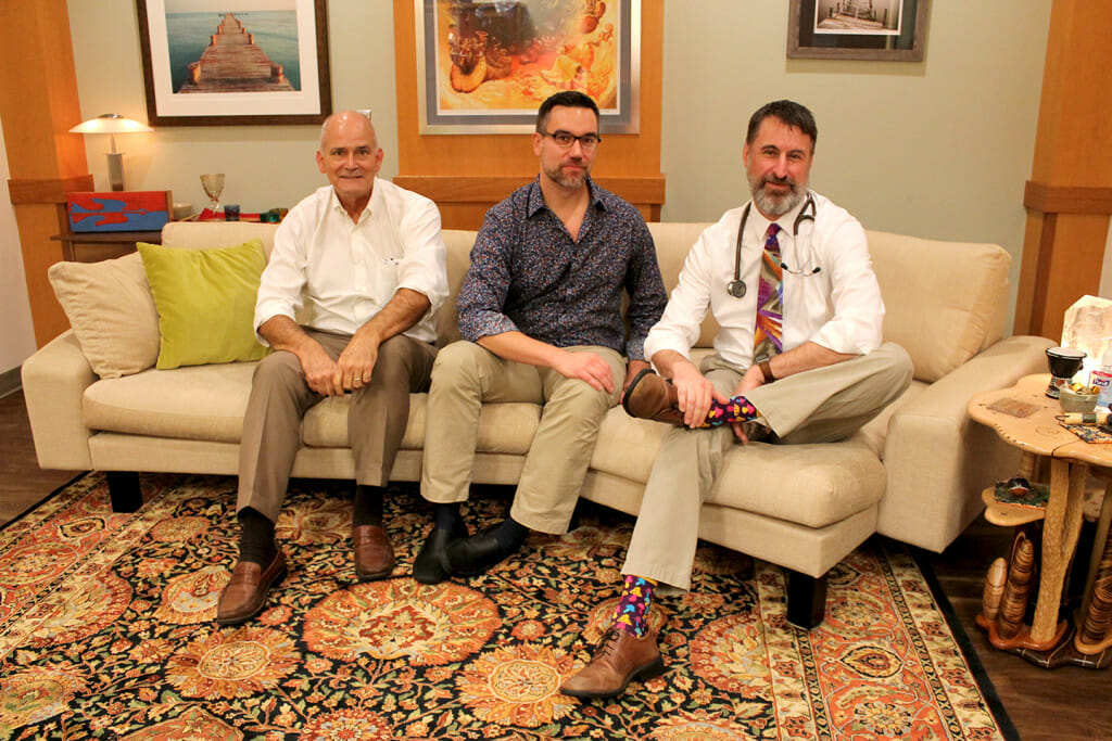 School of Pharmacy Professor Paul Hutson (left), with School of Medicine and Public Health collaborators Assistant Professor Christopher Nicholas (center) and Professor Randall Brown (right) sit on a cream sofa and smile toward the camera. They are in the room used for psychedelic testing.