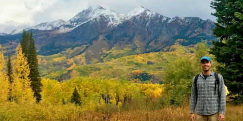 Ford Freyberg appears small in the lower righthand corner of the photo as he stands in front of the Rocky Mountain range on an autumn day. The forest in the foreground is changing from green to gold.