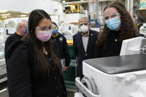 Two women in surgical masks and business casual attire look at a monitor next to a bench-top piece of lab equipment shaped like a cooler. Three people in similar attire stand in the background. They are in an industrial setting. The room appears to be filled with equipment.