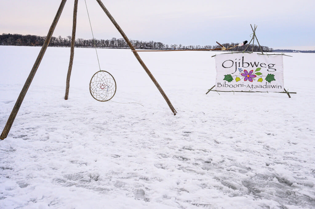 A hoop for the hoop and spear game (dakobijigan-minawaa zhiimaagan) and a sign welcome visitors to the Ojibwe Winter Games on Lake Mendota.