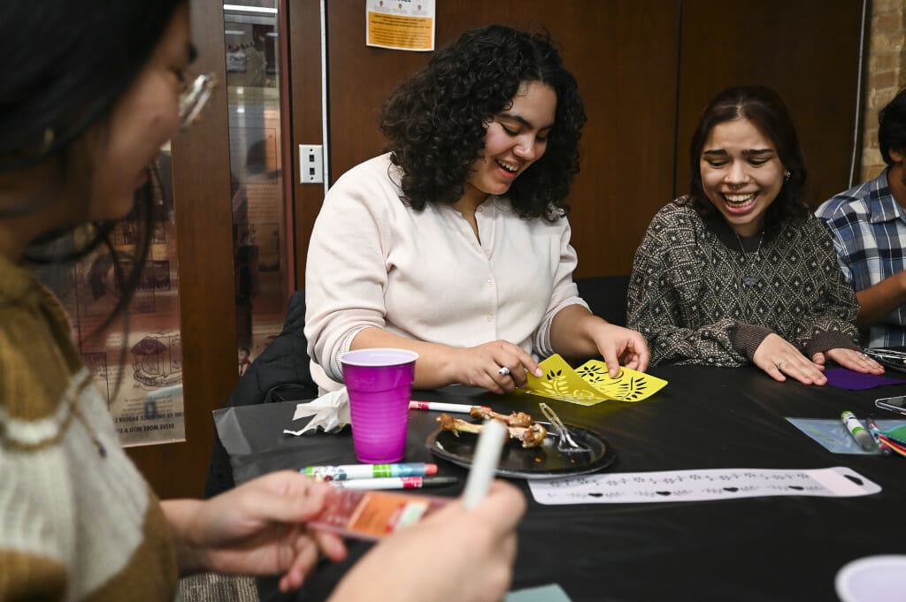 Sophia Whitehead, left, and Jasmine Lutz, right, laugh while decorating textiles. They are seated around a table covered in fabric squares, stencils and markers.
