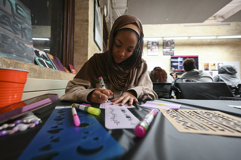 Seated at a table, Sakinah Yusuf leans in to decorate a fabric square using a stencil and markers.