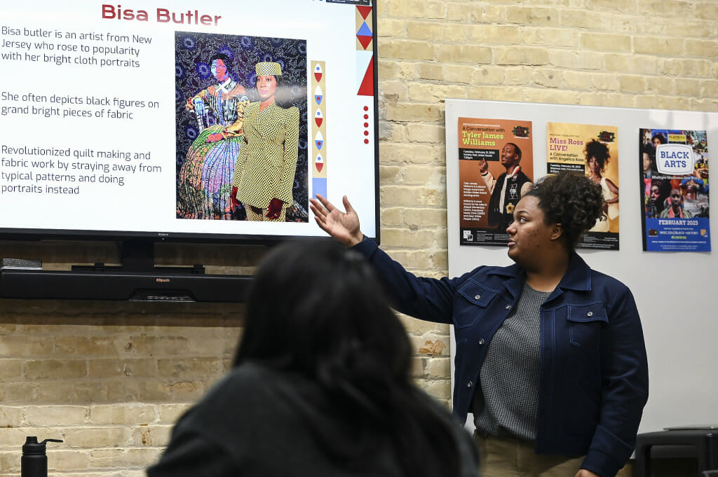 Jasmine Jones stands and gestures toward a screen displaying photos of the colorful quilting work of Bisa Butler.
