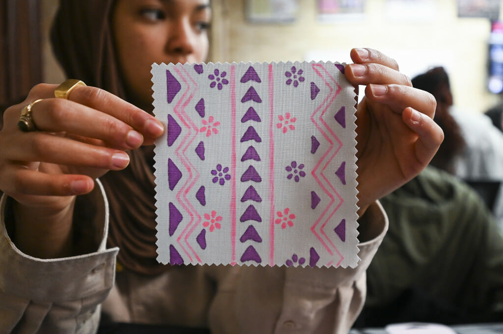 Sakinah Yusuf displays a decorated textile that was created with markers and will later be made into one collective quilt as part of the ‘Quilt Together. Stay Together’ event. She has decorated the fabric square in geometric and floral patterns using pink and purple markers.