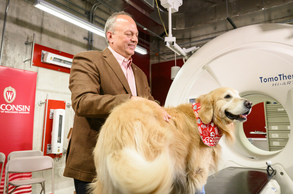 Scout, a golden retriever stands on an exam table and an medical imaging device. He is wearing a red and white bandana printed with Bucky Badger, the Wisconsin W and his name. His owner, WeatherTech Founder and CEO David MacNeil, stands at his side smiling. 