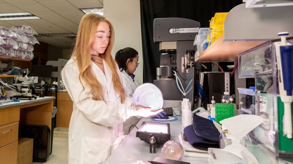 A woman with long blond hair and a white lab coat holds a plastic sphere over a lab bench. Another researcher works at a machine behind her.