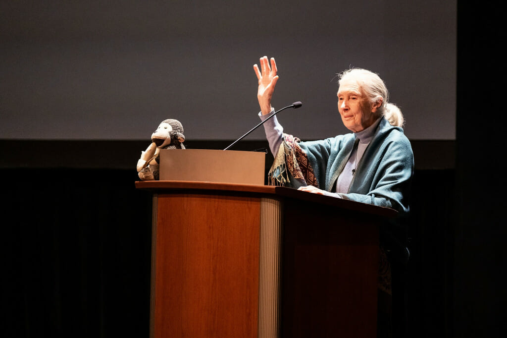 A woman at a podium waves to the audience.