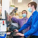 Behind the counter in a pharmacy, two men wearing button-down shirts and slacks stand in front of a computer. They are both wearing blue surgical masks. One man points to information on the computer monitor while the other types on the keyboard.