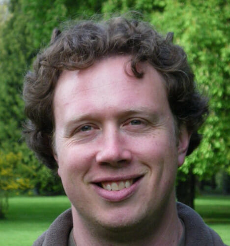 A headshot of Olaf Jensen, shot outdoors against a green foliage background. Jensen has short brown curly hair and is smiling toward the camera.