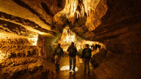 A well-lit cave with stalagmites hanging down. A group of three people walk away from the camera through an open passageway.