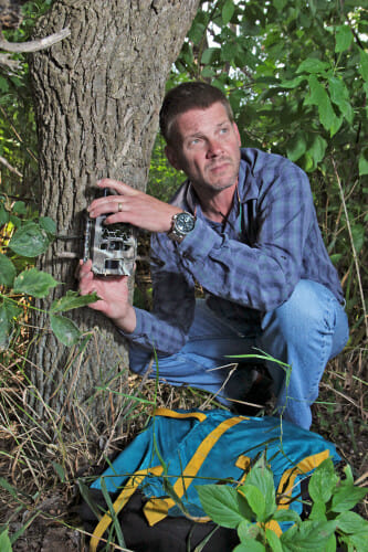 Tim Van Deelen crouches by a tree trunk in the woods. He is positioning a trail camera on the tree and looking ahead down the trail camera's sight line.