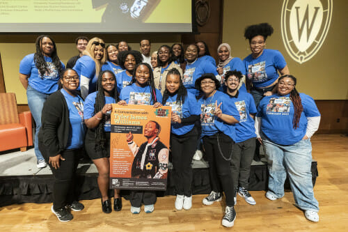 A photo of the 2023 Black History Month planning committee standing in an auditorium. They're wearing matching blue shirts and holding a poster for their keynote event.