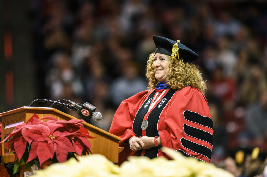 UW–Madison Chancellor Jennifer L. Mnookin stands at a podium in front of a large crowd in the Kohl Center. She is wearing academic regalia in red and black.