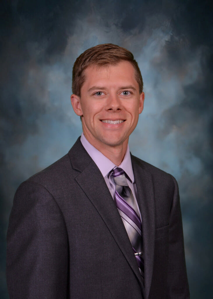 A headshot portrait of Nicholas Hess. Hess has short, dark blond hair. He is smiling to the camera. He is wearing a charcoal gray suit with a light purple shirt and tie. He is standing against a studio backdrop.