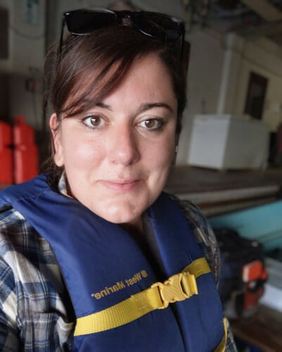 Grace Wilkinson has brown hair with side-swept bangs. She is wearing a blue life vest over a plaid shirt and smiling to the camera.