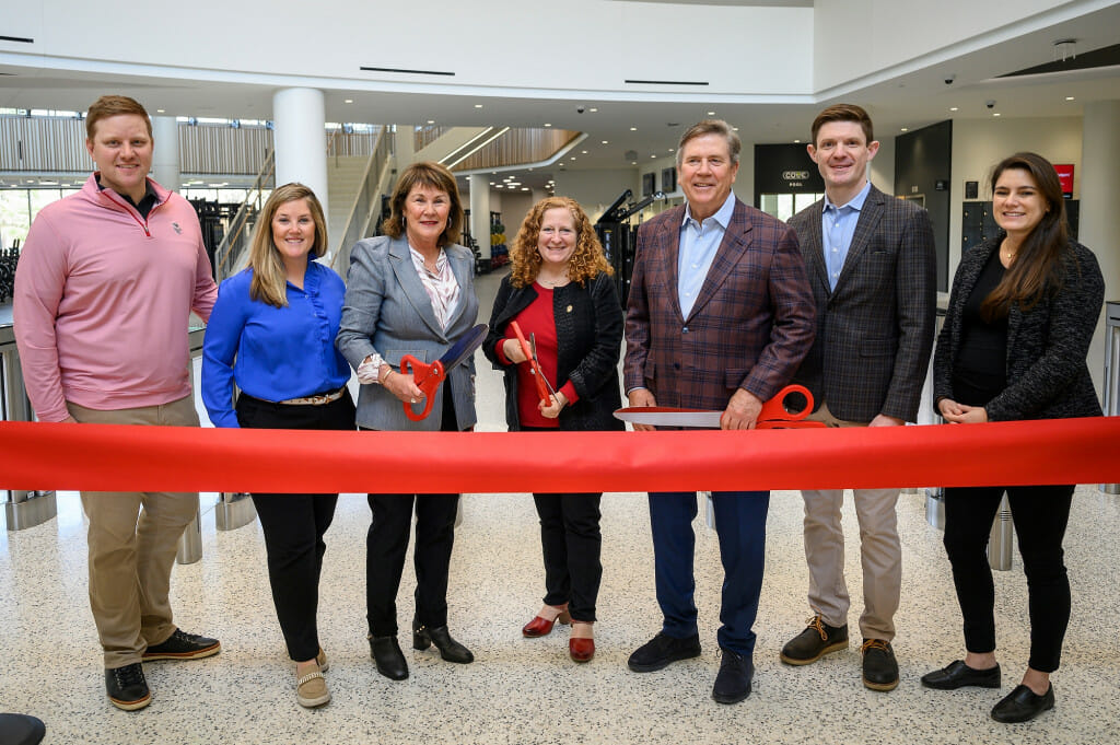 A group of people stand by a big red ribbon, ready to cut it with scissors.