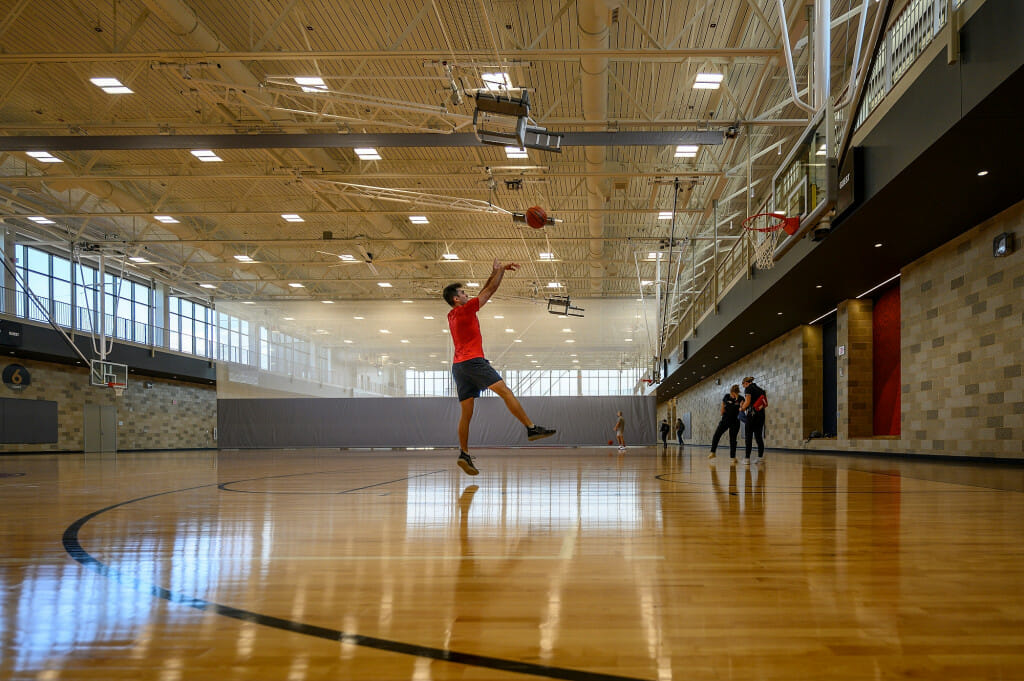 A man shoots a basket in a vast basketball court, with a shiny new hardwood floor.