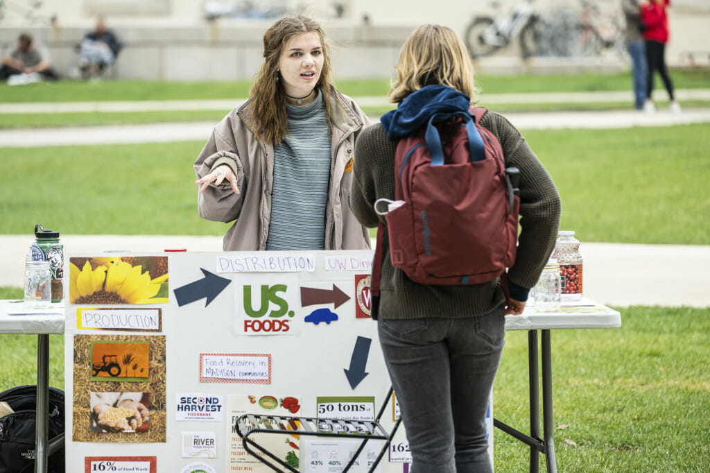 Two women talk over a display table featuring a poster about minimizing food waste and food insecurity. They are outdoors on Library Mall on a chilly spring day.