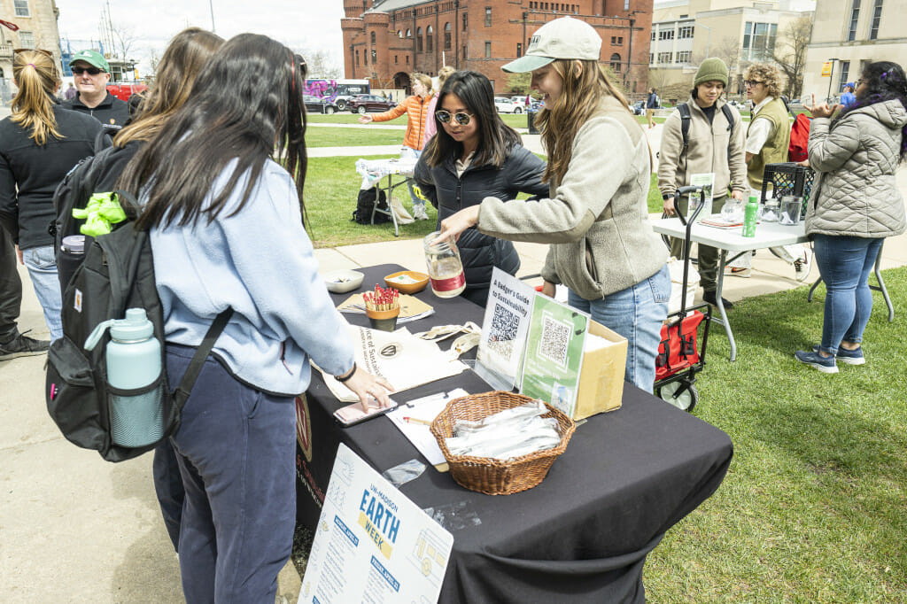 A group of four women gather at a display table to talk about themes related to Earth Day. They are outdoors on Library Mall on a chilly spring day.