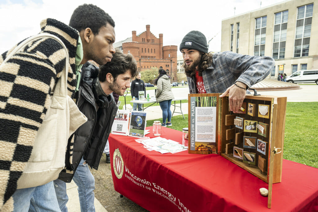 Two men lean over a table to look at a display. A third man stands behind the display as he speaks to the visitors at his table. They are outdoors on Library Mall on a chilly spring day.