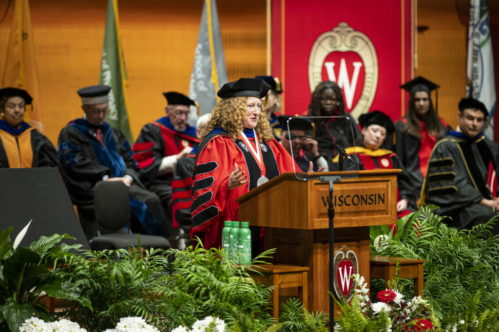 Jennifer Mnookin stands on stage with a crowd of dignataries, all wearing academic regalia. Mnookin folds her hands on her heart in a gesture of gratitude as she makes an address from a wooden podium with the word 