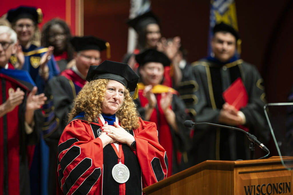 Jennifer Mnookin stands on stage with a crowd of dignataries, all wearing academic regalia. Mnookin folds her hands on her heart in a gesture of gratitude as she makes an address from a wooden podium with the word "Wisconsin" and the UW-Madison crest on the front.