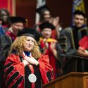 Jennifer Mnookin stands on stage with a crowd of dignataries, all wearing academic regalia. Mnookin folds her hands on her heart in a gesture of gratitude as she makes an address from a wooden podium with the word 