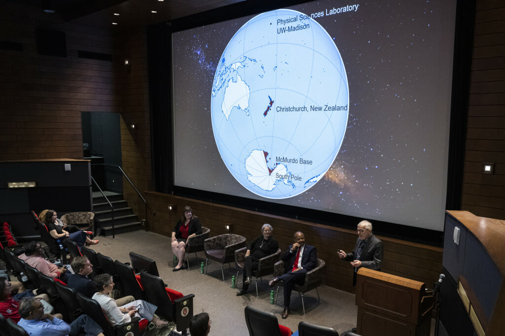 Professor Francis Halzen stands at the front of an auditorium and speaks to a crowd. On the screen is a map showing where the IceCube observatory is located. 