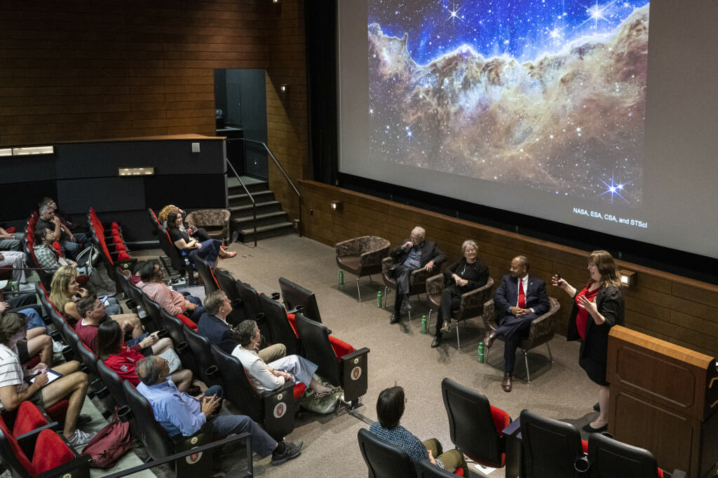 A lecture hall full of people look at an image of clouds on a screen.