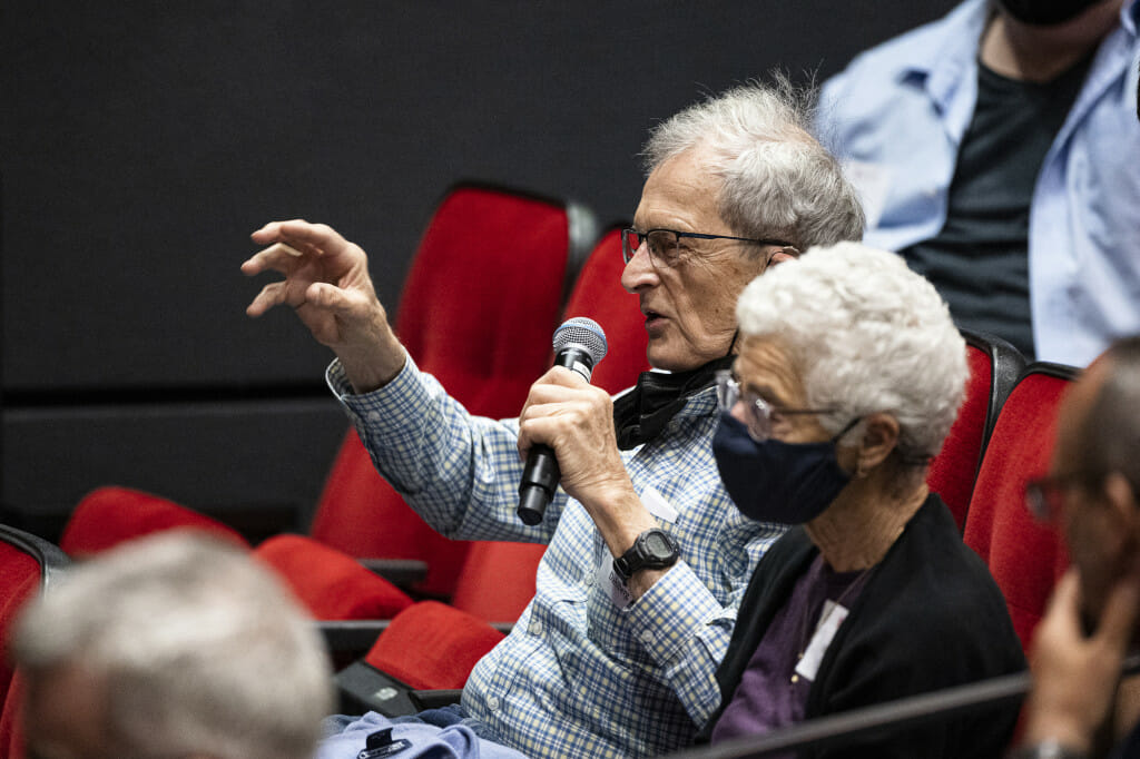 A man sitting in the audience holds a microphone and gesticulates as he asks a question.