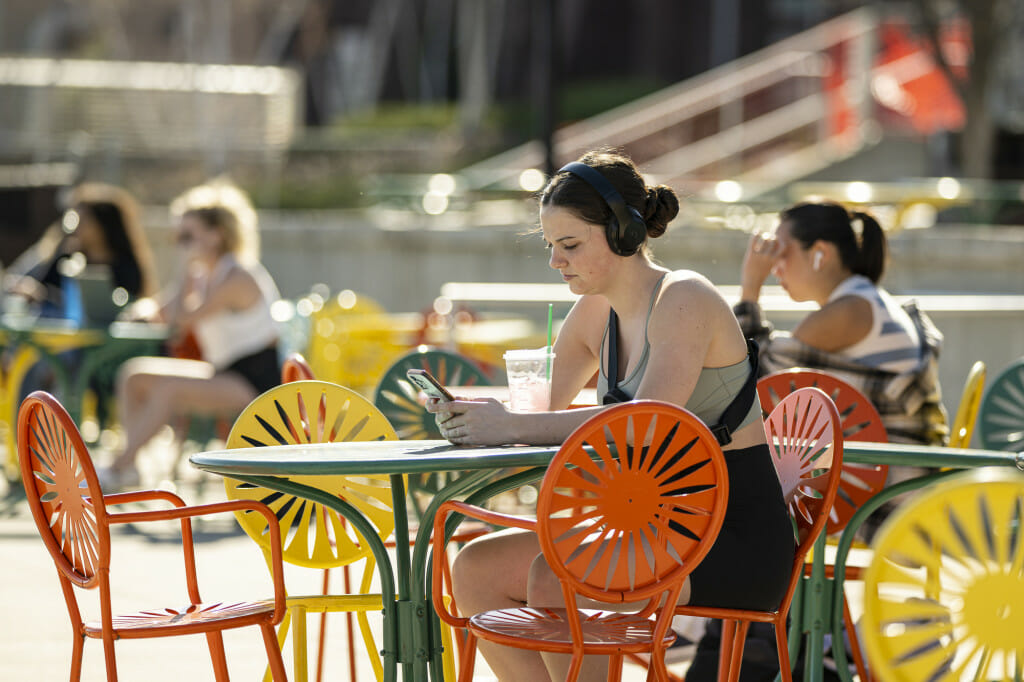 A student, Sami Lacke, wears headphones and looks at her phone while sitting at a table at the Memorial Union Terrace on a sunny day.
