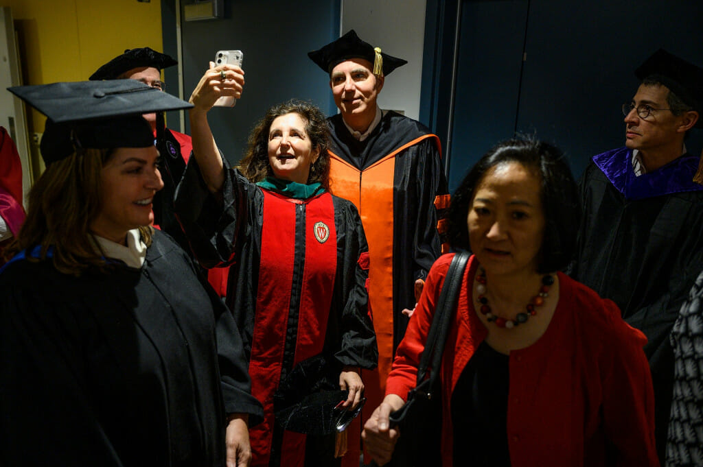 A tight shot of six people standing in a corridor wearing academic robes and hats. A woman at center raises her phone to take a selfie with another member of the group.