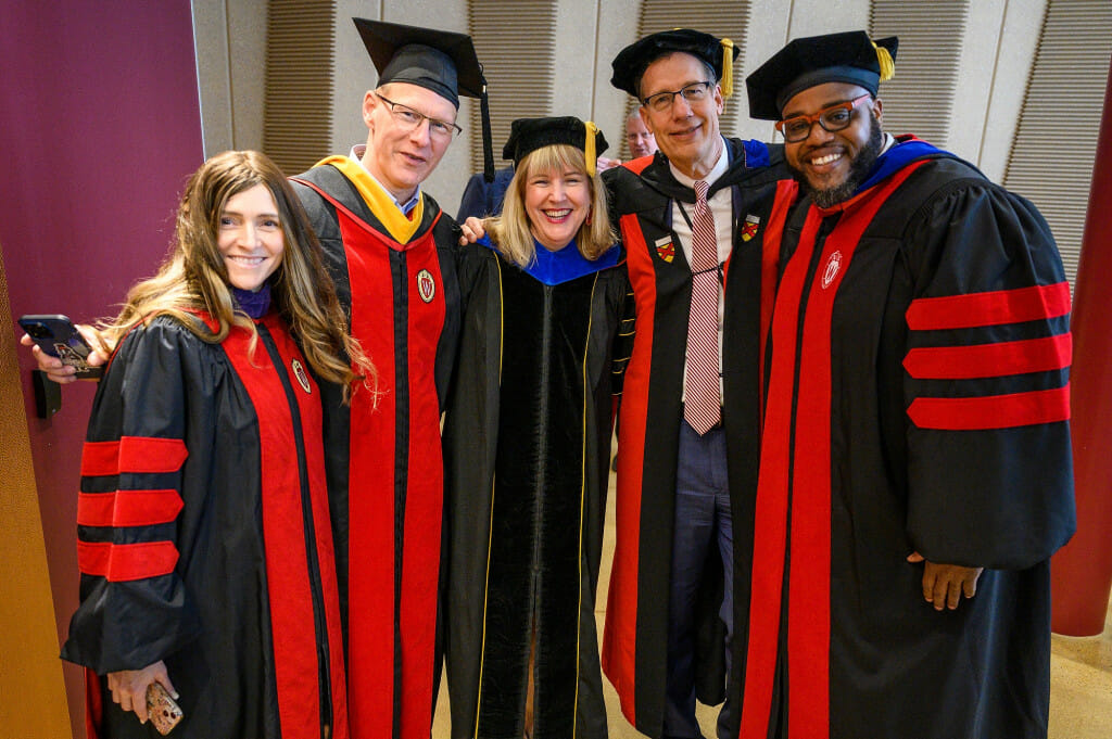 Five people stand in a row with their arms around each other's shoulders and smiling at the camera. They are wearing academic robes and hats