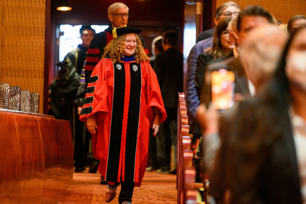 Jennifer Mnookin, wearing red academic robes and a black doctoral hat, walks into an auditorium. Governor Tony Evers, also wearing academic regalia, walks behind her.