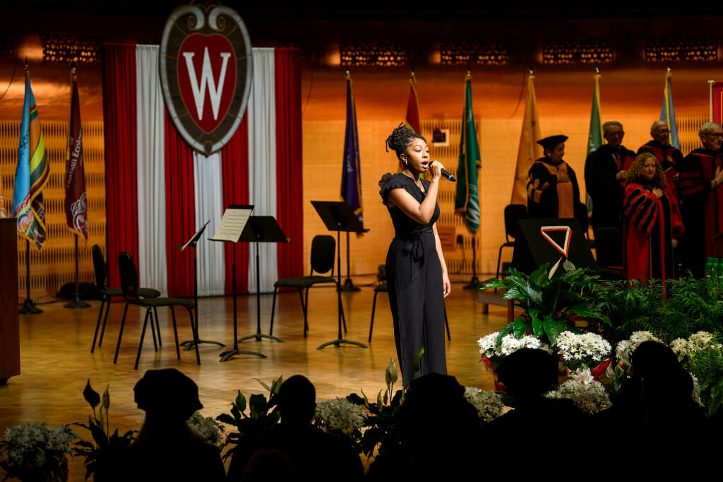 A woman sings the national anthem on stage in front of the UW–Madison crest. 