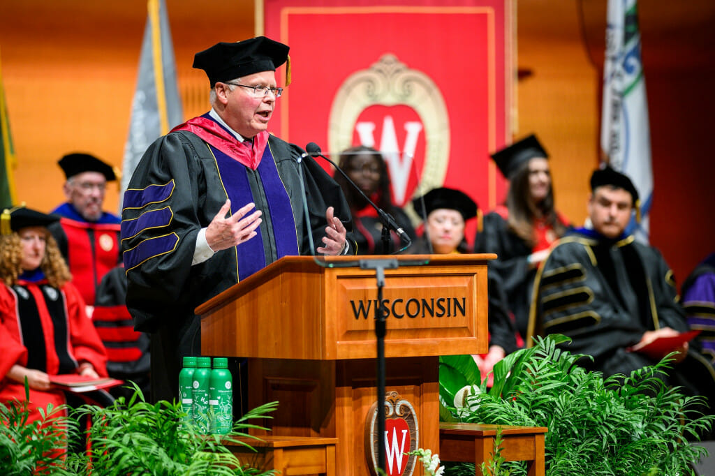 A man wearing doctoral robes and hat speaks at a lectern.