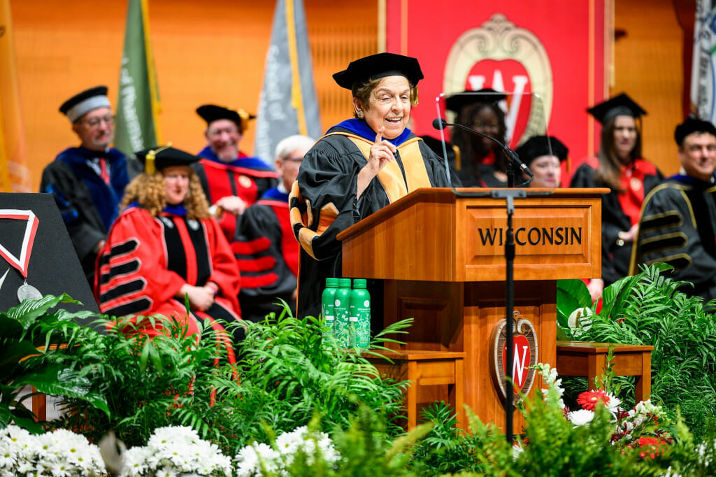 A woman wearing doctoral robes and hat speaks at a lectern.