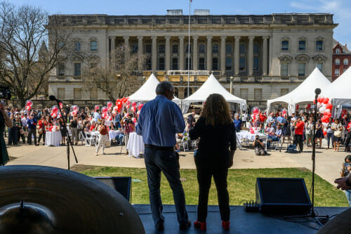 Two people stand on a stage in front of a sunny plaza, addressing the people there.