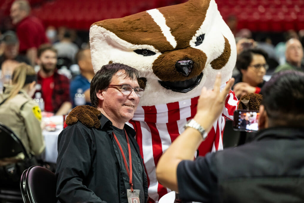 Jason Behlke smiles and poses for a photo with Bucky Badger amidst a crowd seated at banquet tables on the floor of the Kohl Center arena.