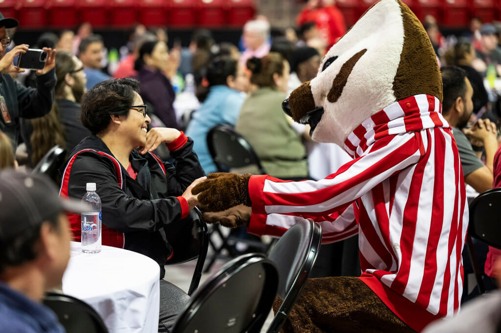 Estela Ramirez smiles and holds hands with Bucky Badger amidst a crowd seated at banquet tables on the floor of the Kohl Center arena.