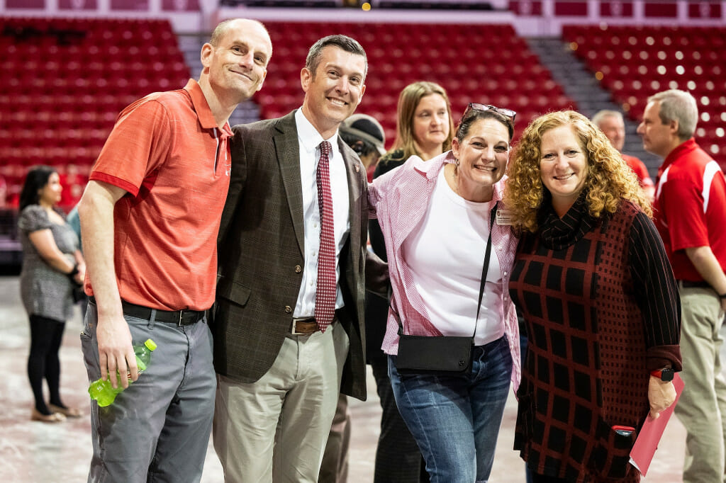 A group of four people, Paul Seitz, Patrick Sheehan, Cindy Torstveit and Jennifer Mnookin stand with their arms around each other's shoulders and smile at the camera. They are standing on the floor of the Kohl Center arena.
