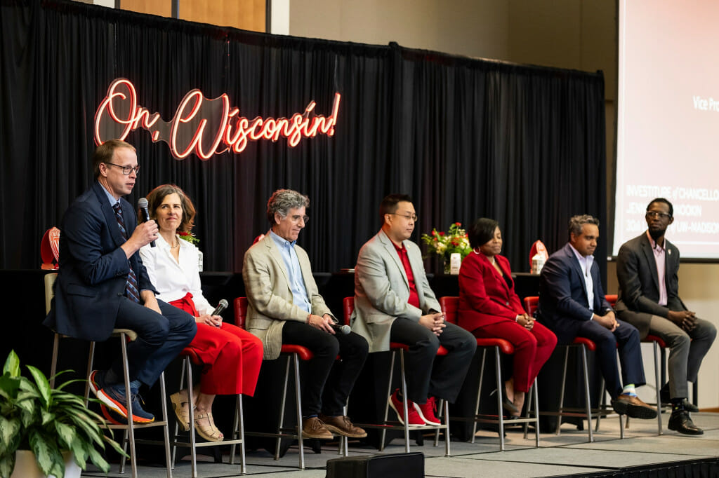 Seven people in a row sit on stools on a stage with a black backdrop and a neon sign reading "On, Wisconsin!" behind them